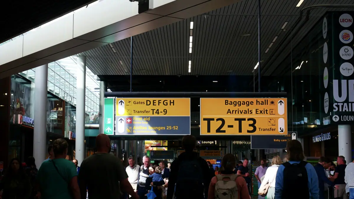 Travellers walking below the yellow arrivals and baggage hall signs inside Schiphol Airport